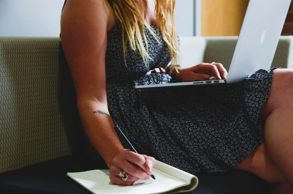 Femme assise sur un canapé, prenant des notes sur un carnet tout en utilisant un logiciel de facturation gratuit sur son ordinateur portable